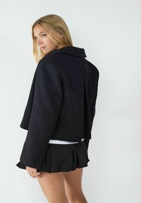 Schwarze cropped Jacke mit breitem Kragen und langen Ärmeln, aus einem glatten Material, kombiniert mit einem gefalteten schwarzen Rock.