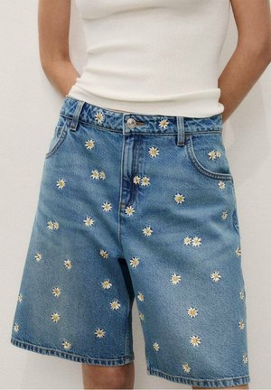 Short en denim avec broderie de marguerites blanches et jaunes, porté avec un top blanc sans manches côtelé.