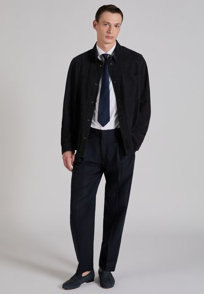 Młody mężczyzna w czarnej kurtce, białej koszuli, granatowym krawacie, granatowych spodniach i ciemnych mokasynach stoi na szarym tle, mając jedną rękę w kieszeni.