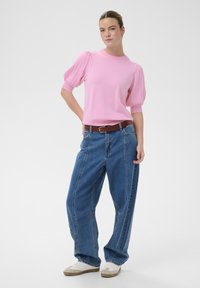 Žena stojící s rukama v bok, oblečená v růžovém topu s nadýchanými rukávy, volných modrých džínách s hnědým pásem a bílými teniskami na bílém pozadí.