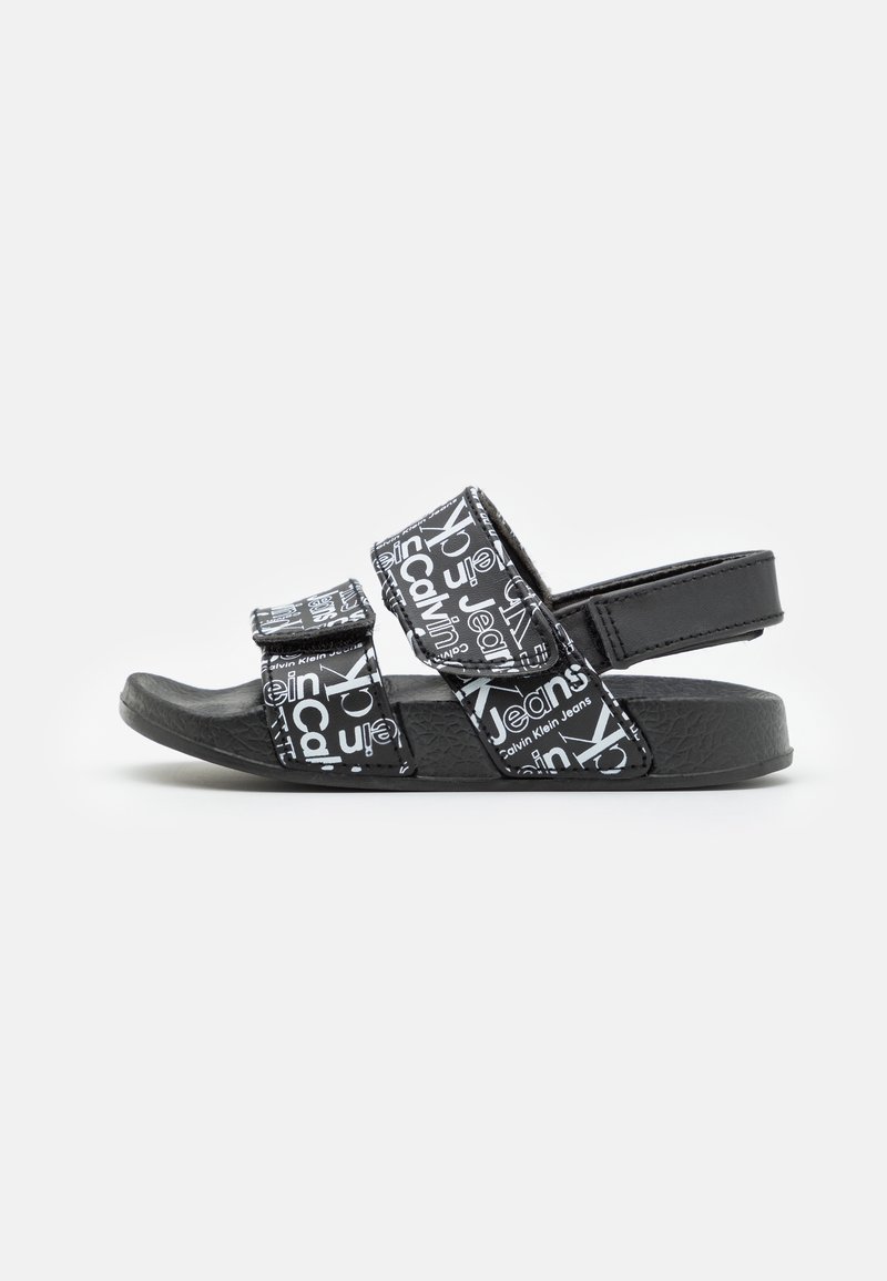 Černé sandály se dvěma nastavitelnými popruhy, které mají bílé logo. Texturovaná podešev a hladké svrchní materiály. Zobrazeno z bočního pohledu.