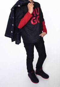 Kind trägt ein schwarz-rotes Lagenhemd mit "HUGO"-Schriftzug, schwarze Jeans, schwarz-rote Sneakers und hält eine schwarze Jacke über der Schulter.
