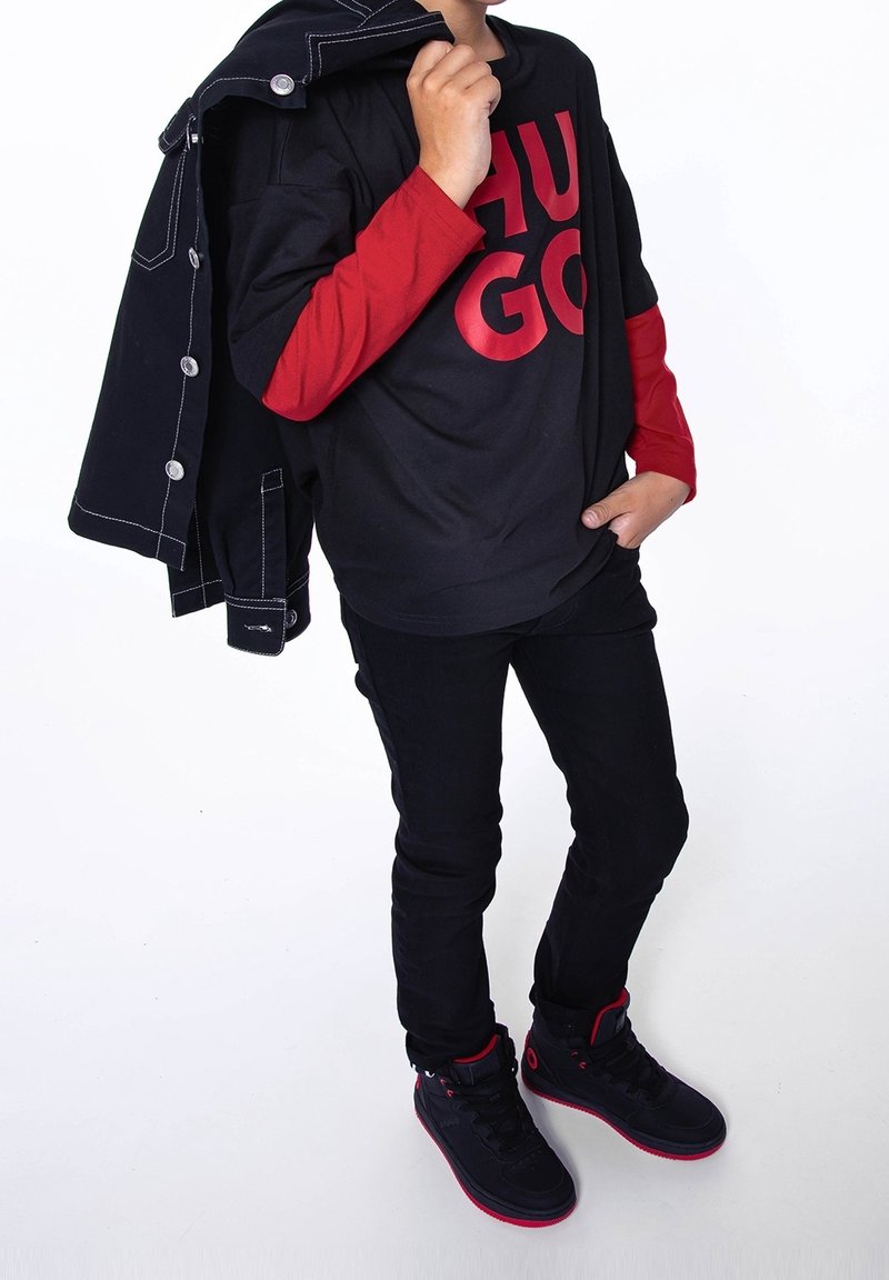 Kind trägt ein schwarz-rotes Lagenhemd mit "HUGO"-Schriftzug, schwarze Jeans, schwarz-rote Sneakers und hält eine schwarze Jacke über der Schulter.