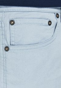 Světle modrá denimová tkanina zblízka, kde se nachází přední kapsa s měděnými nýty a kontrastním prošíváním.