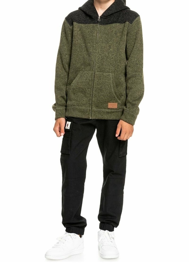 Sudadera con cremallera color verde oliva con un panel de hombro texturizado negro, dos bolsillos frontales y una etiqueta de tela. Combinada con pantalones cargo negros y zapatillas blancas.