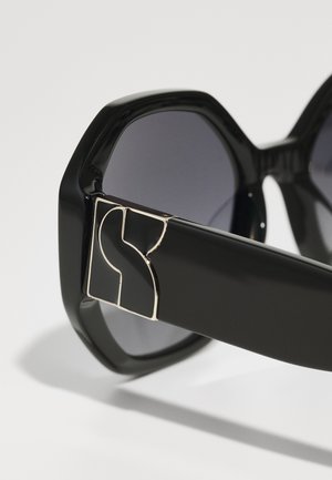 Černé oversized sluneční brýle s hexagonálními čočkami, lesklým rámečkem a zlatým logem na spánku, které předvádějí elegantní a moderní design.