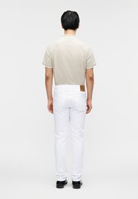 Μπεζ πουκάμισο πόλο με σχέδιο και λευκή διακόσμηση, συνδυασμένο με λευκό τζιν και μαύρα παπούτσια, από πίσω. Δερμάτινη ετικέτα στη ζώνη μέσης.