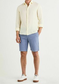 Camisa de lino amarillo claro con botones, mangas remangadas; pantalones cortos azules; zapatillas blancas con suelas marrones. Diseño simple y casual.
