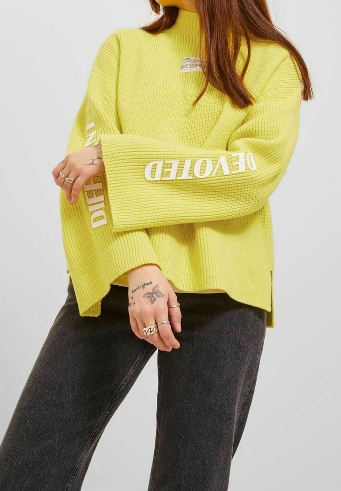 Personne portant un pull jaune côtelé avec du texte blanc sur les manches et un jean noir, montrant une main tatouée avec plusieurs bagues.