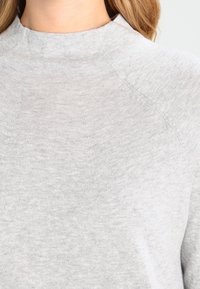 Hellgrauer Strickpullover mit einem dezenten, erhöhten Muster am Halsausschnitt, getragen von einer Person mit langen blonden Haaren.