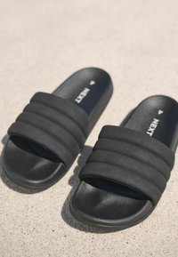 Pár fekete, bebújós papucs párnázott pántokkal, egy texturált világos felületen pihenve.