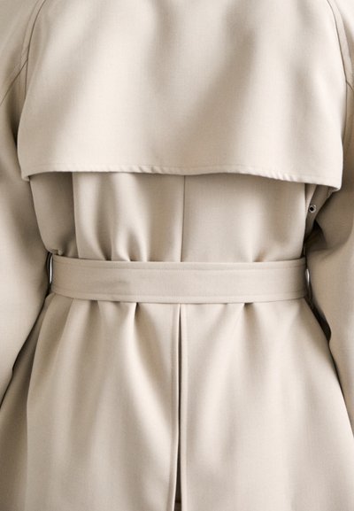 Trench coat beige con ampio collare, vita stretta e pieghe sul retro. Realizzato in tessuto morbido, presenta un design minimalista.