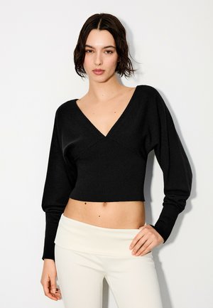 Femme aux cheveux courts et foncés portant un pull court noir à manches longues et col en V ainsi qu’un pantalon blanc taille haute, debout devant un fond blanc.