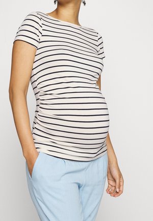 Personne enceinte portant une chemise blanche à rayures horizontales noires et un pantalon bleu clair, debout avec une main dans la poche.