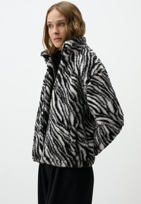 Fluffy zwart-wit gestreepte zebra-jack met hoge kraag, ritssluiting en een relaxed fit. Zachte textuur, ontworpen voor warmte en comfort.