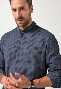 Koszula z długim rękawem z dżerseju w odcieniu granatu, z geometrycznym wzorem. Posiada kołnierzyk z ostrym zakończeniem, jedną kieszeń na piersi oraz zaokrąglone mankiety.