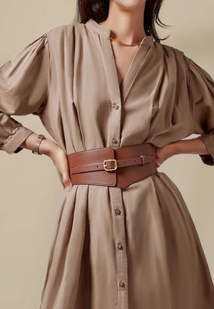Brown leather belt with a rectangular shape and gold buckle, worn over a beige, button-up, textured dress with puffed sleeves.
