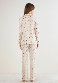 Kremowy komplet piżamy z długimi rękawami i spodniami, zdobiony wzorem czerwonych kokardek i neutralnych kształtów, z kontrastowymi lamówkami.