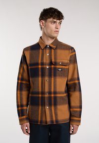 Camicia a quadri marrone e blu navy con colletto a punta, chiusura a bottoni, tasca sul petto e bottoni metallici, realizzata in un tessuto testurizzato.