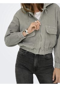 Krótkia, jasno szara kurtka sztruksowa z kapturem, zapinana z przodu na zamek błyskawiczny, z dwoma kieszeniami na piersiach, w zestawie z ciemnymi dżinsami. Gładka, teksturowana tkanina.