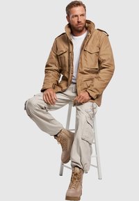 Chaqueta militar color caqui con dos bolsillos en el pecho, llevada sobre una camiseta blanca. Combinada con pantalones cargo de color beige claro y botas de encaje color caqui.
