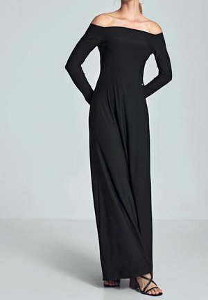 Mujer que lleva un vestido largo negro con los hombros descubiertos y mangas largas, y sandalias negras de tacón con tiras, de pie con las manos detrás de la espalda.