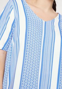 Nainen, jolla on yllään valkoisista ja sinisistä raitoista tehty lyhythihainen paita, jossa on V-pääntie. Paidassa on monimutkaisia geometrisia kuvioita sinisissä raidoissa.