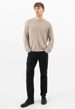 Jonge man staand met handen in de zakken, gekleed in een beige gebreide trui, zwarte broek en zwarte veterschoenen tegen een witte achtergrond.