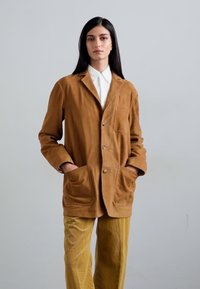 Blazer in suede marrone con due tasche frontali, chiusura a singolo bottone e colletto classico. Abbinato a pantaloni gialli in corduroy.