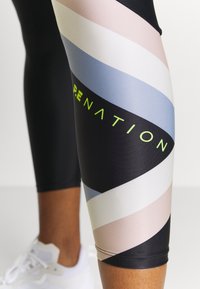 Zbliżenie na nogę w czarnych legginsach z pastelowymi paskami i neonowymi żółtymi napisami, połączoną z białymi butami sportowymi.