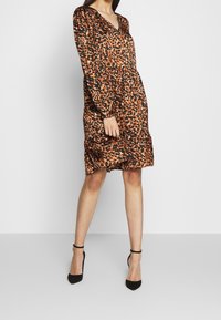 Leopardmönstrad klänning i brunt, svart och orange. Långa ärmar, V-hals, figurnära midja, lätt utsvängd fåll. Slät yta, knälång skärning.