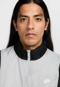 Αντιανεμικό μπουφάν Nike από ελαφρύ, γκρι ύφασμα με μαύρο γιακά. Διαθέτει φερμουάρ μπροστά και μικρό λευκό λογότυπο στο στήθος.