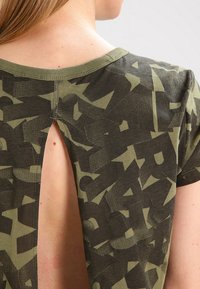 Olivově zelené tričko s číselným potiskem, měkkou texturou a výřezem na zádech. Kolo výstřihu a krátké rukávy.