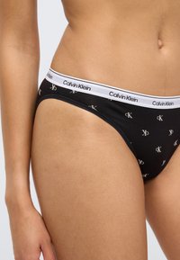 Μαύρο βαμβακερό μπικίνι εσώρουχο με λευκή ταινία που φέρει το "Calvin Klein." Το ύφασμα έχει ομαλή υφή και επαναλαμβανόμενο μοτίβο λογότυπου.
