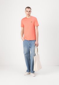 Koralfärgad t-shirt med rund halsringning, korta ärmar, liten gul logotyp, kombinerad med ljusblå vida jeans och en beige canvas toteväska. Vita sneakers.