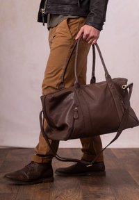 Brown leather duffel bag with dual handles, side zipper pocket, and removable shoulder strap. Smooth texture, spacious design, and minimal accents.