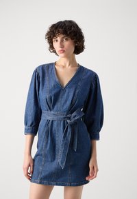 Denimkjole med dyp V-hals, boblede lange ermer og belte i stoff i livet. Lys blå farge med kontraststing.
