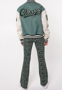 Green and cream varsity jacket with "Chicago" text, paired with flared green floral pants. White sneakers complete the outfit.