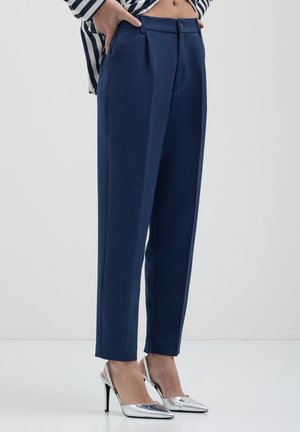 Marineblaue maßgeschneiderte Hose mit glatter Textur, mit Falten, einem Knopfverschluss und seitlichen Taschen, kombiniert mit silbernen High Heels.