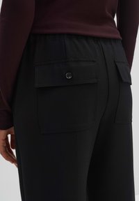 Un pantalon cargo noir présente une coupe décontractée, une taille élastique et deux poches arrière à boutons. Le tissu semble lisse avec un éclat minimal.