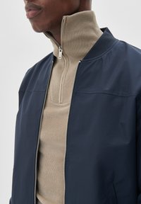 Marineblauwe bomberjack over een beige geribbelde coltrui. Het jack heeft een hoge kraag en een voorrits met zilverkleurige hardware.