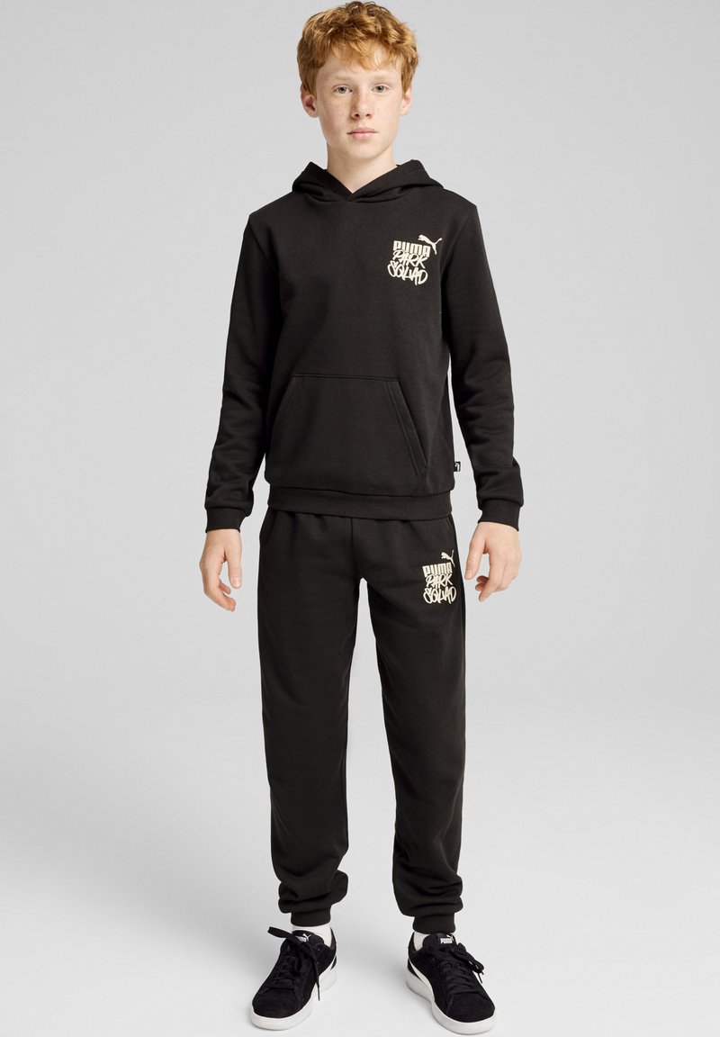 Conjunto de sudadera con capucha y pantalones de chándal de tejido suave. La sudadera presenta un bolsillo frontal y un detalle de logo. El modelo lleva zapatillas negras.