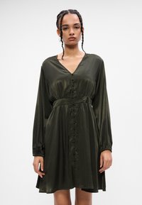 Σκούρο πράσινο σατέν φόρεμα με V λαιμόκοψη, μακριά μανίκια και κουμπιά μπροστά. Διαθέτει συγκεντρωμένη μέση και φουσκωτό φούστα. Λείος υφασμα.