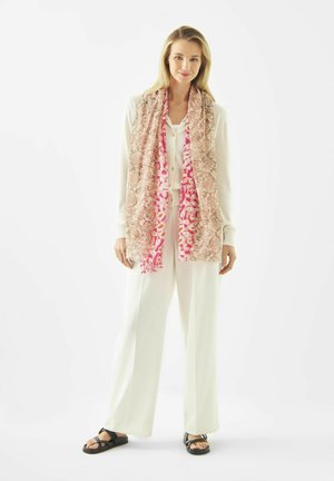 Vrouw staande, gekleed in witte outfit, met een roze sjaal met patroon en zwarte sandalen tegen een effen witte achtergrond.