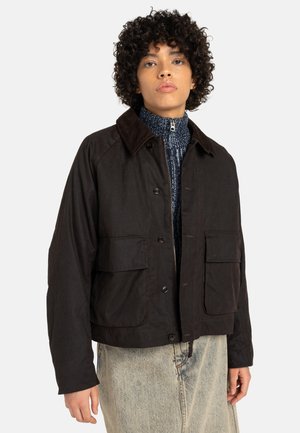 Bruine cropped jas met een corduroy kraag, twee voorzakken en knoopsluiting. Draagt over een gestructureerde blauwe trui en lichte jeans.