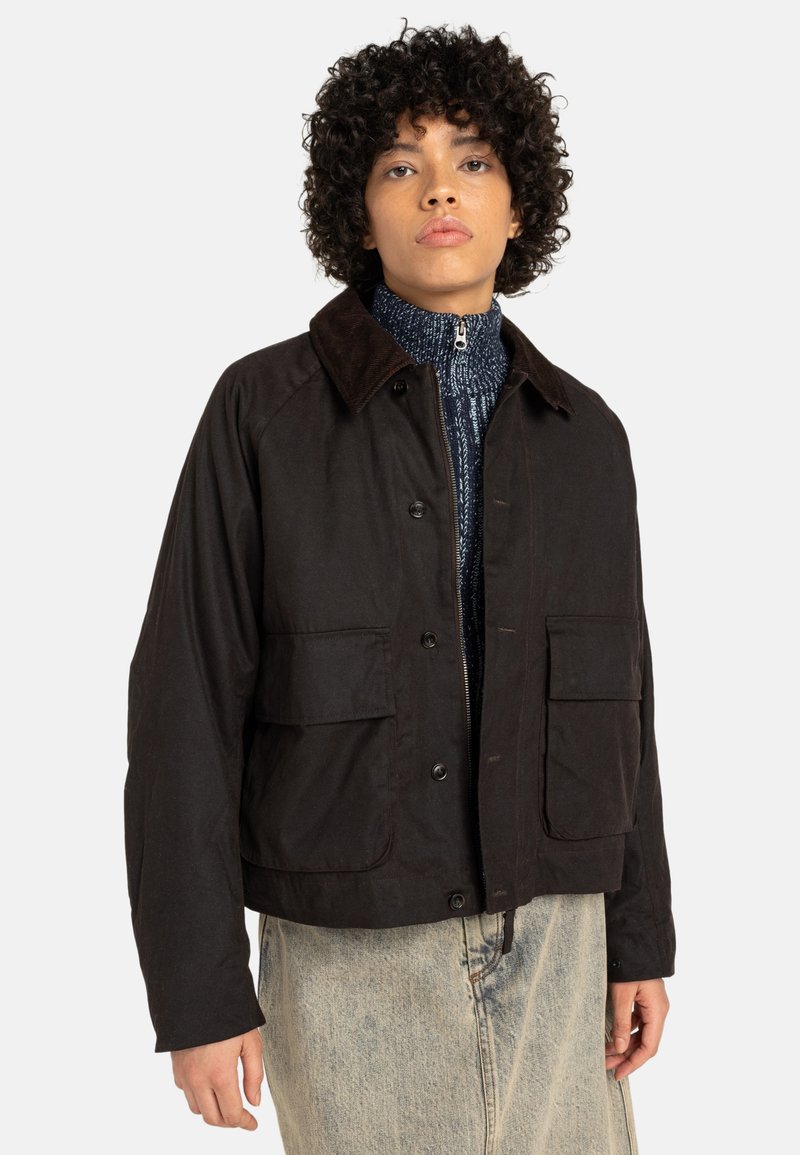 Veste brune courte avec un col en velours côtelé, deux poches avant et une fermeture à boutons. Portée sur un pull bleu texturé et un jean clair.