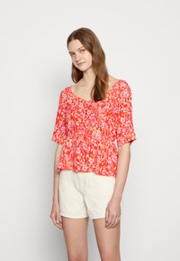 Blusa floral vermelha com mangas curtas, decote arredondado e detalhes franzidos, combinada com calções brancos. Tecido de algodão leve e brilhante.