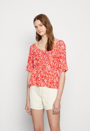 Blusa floral vermelha com mangas curtas, decote arredondado e detalhes franzidos, combinada com calções brancos. Tecido de algodão leve e brilhante.