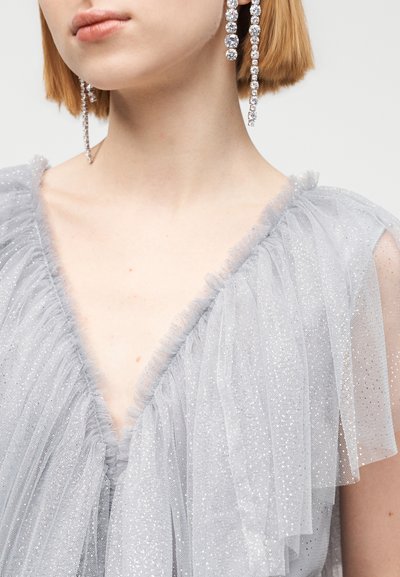 Γκρι φόρεμα από τούλι με κυματιστά τελειώματα, διακοσμημένο με λαμπερά στοιχεία. Βαθιά V-λαιμόκοψη, διάφανα μανίκια και στρωμένη υφή που δημιουργούν οπτικό ενδιαφέρον.