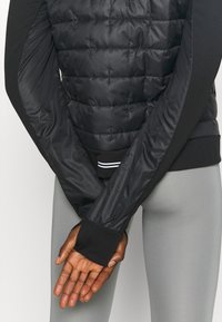 Chaqueta de plumas negra con textura acolchada, que cuenta con mangas largas y puños acanalados. El forro interior es suave y brillante, creando un contraste.